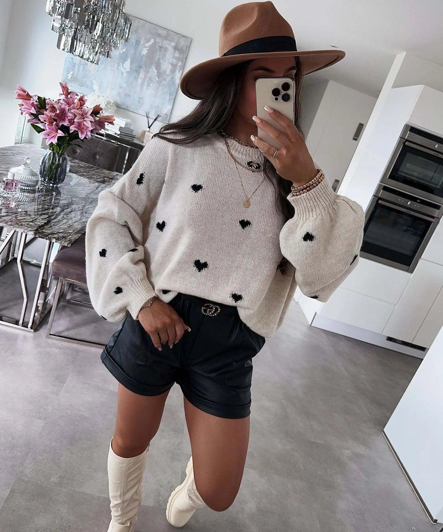 Woman in cozy heart-pattern sweater, black shorts, white boots, and wide-brim hat for stylish comfort wear