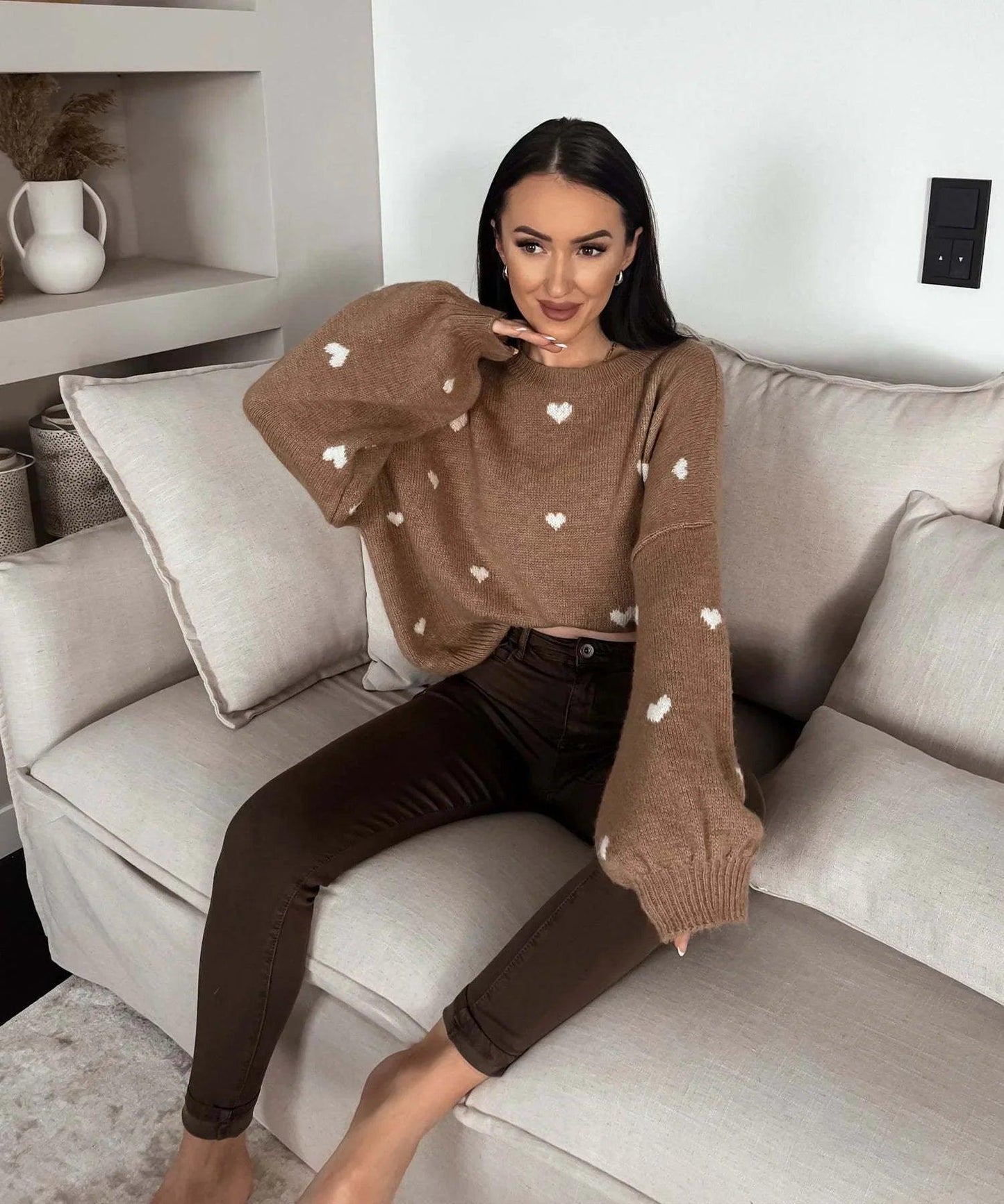 Woman in cozy brown heart-print sweater and dark jeans, sitting on a beige sofa for comfortable everyday style.