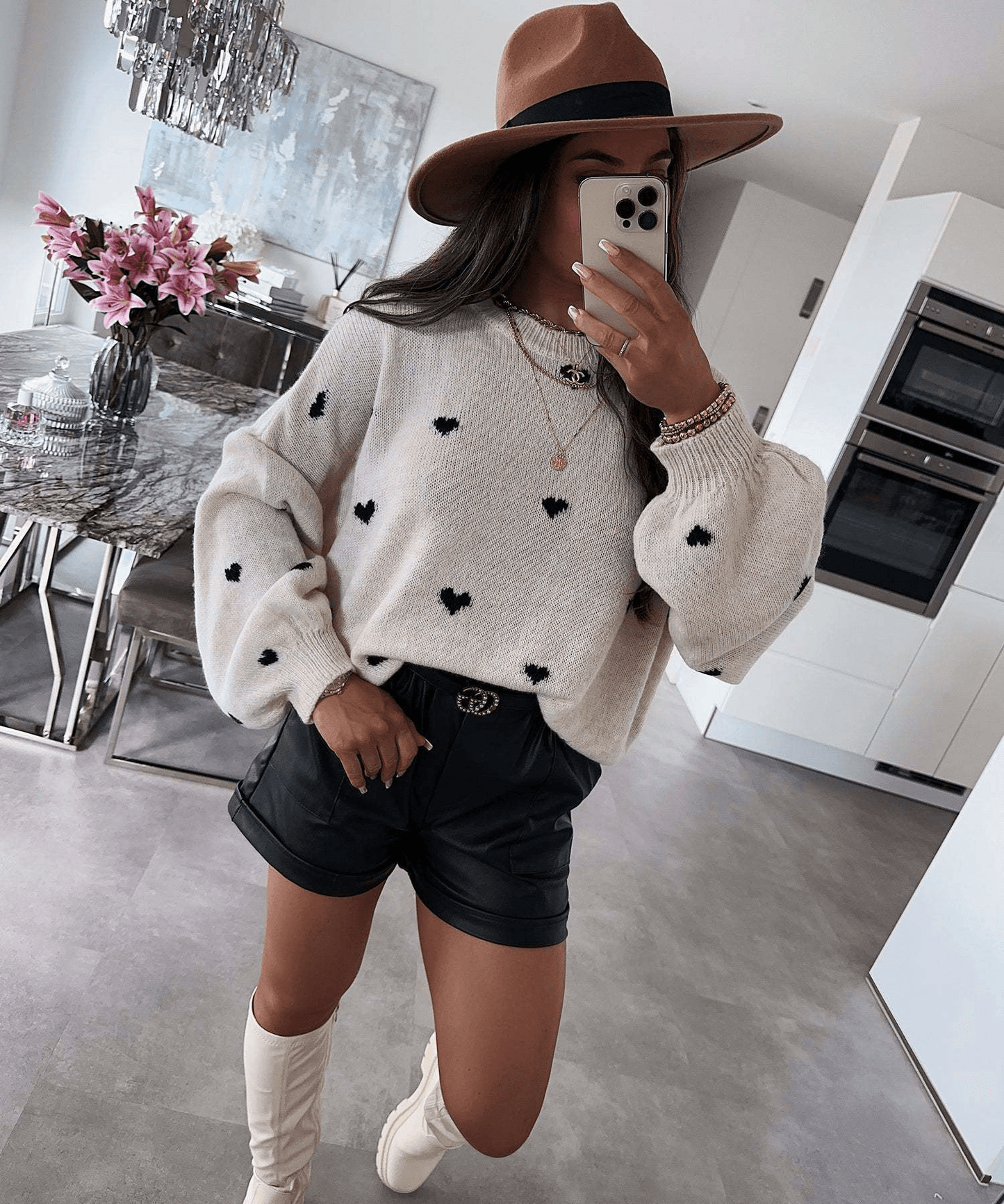 Woman in cozy heart-print sweater, black shorts, white boots, and brown hat for stylish everyday comfort.
